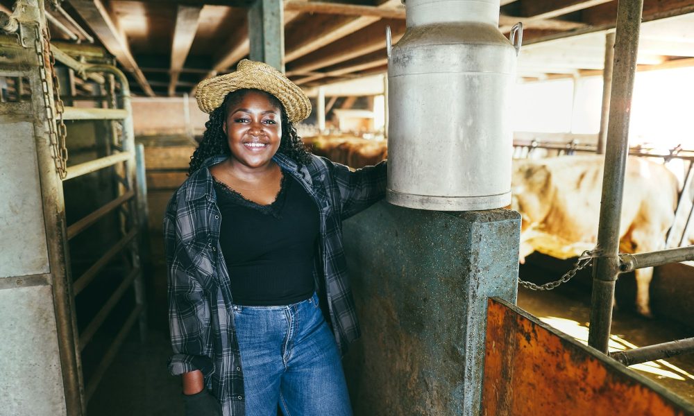 Young african farmer woman holding milk churn inside cowshed - Focus on face
